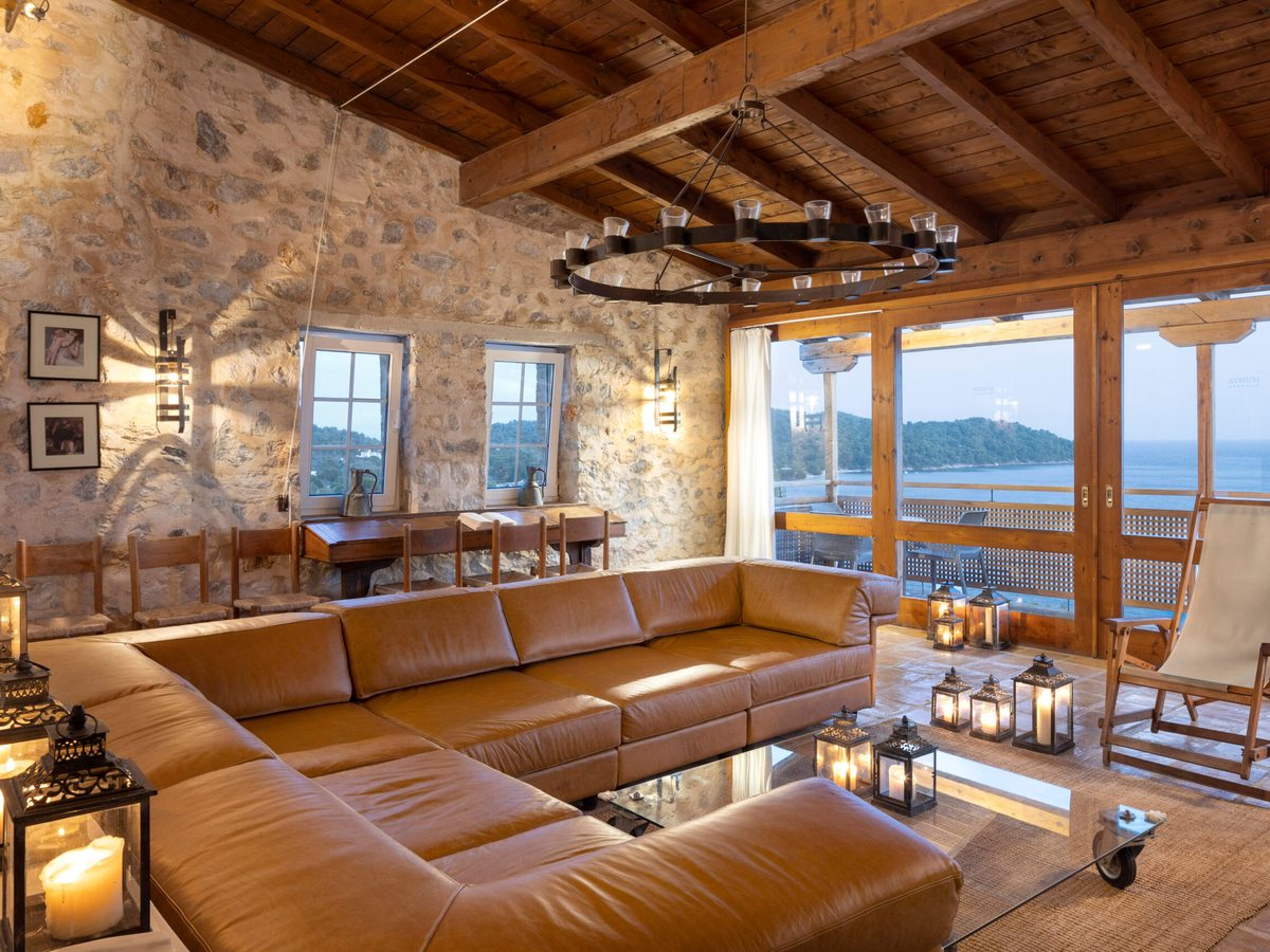 An indoor lounge with exposed stone walls and a timber-beamed ceiling, a tan corner sofa and a coffee table by balcony doors open to a sea view.