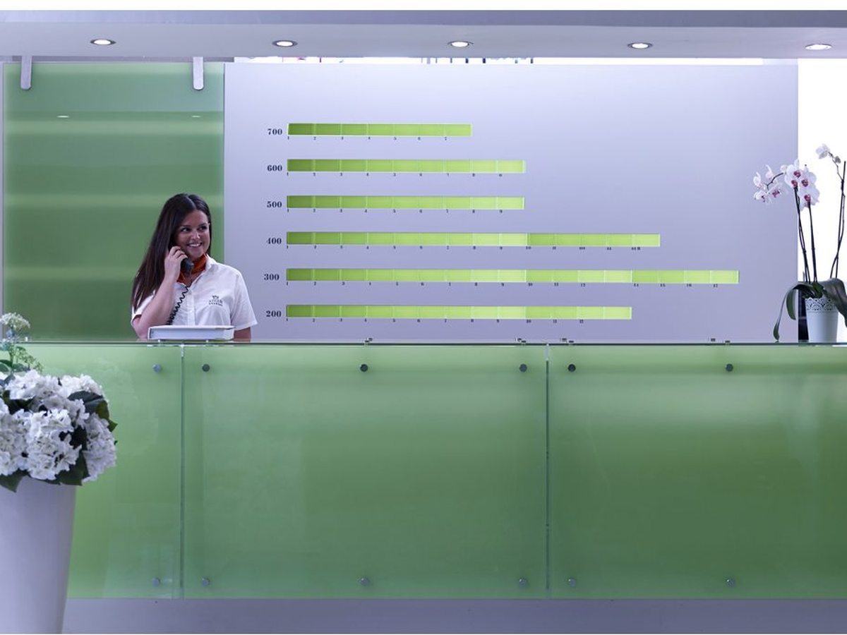 A modern reception desk with a green glass front and a wall display, a staff member behind it and white orchids on the counter.