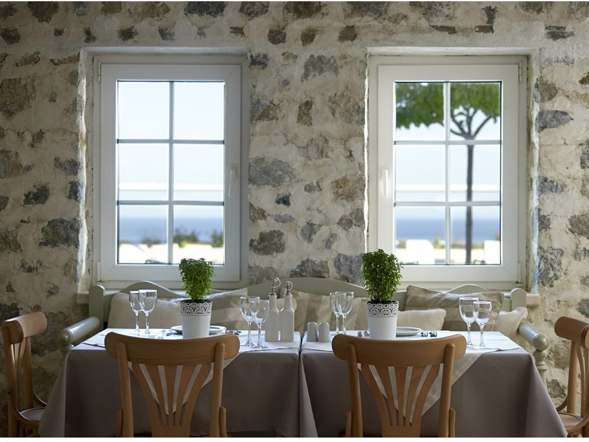 An indoor dining room with exposed stone walls and two white-framed sea-view windows, a set table with wine glasses and potted basil, and wooden bentwood chairs.