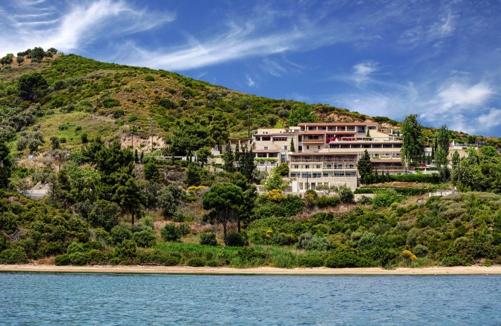 Atrium Hotel Skiathos: stone and wood buildings stepping down a pine-covered hillside above Agia Paraskevi beach and the Aegean.