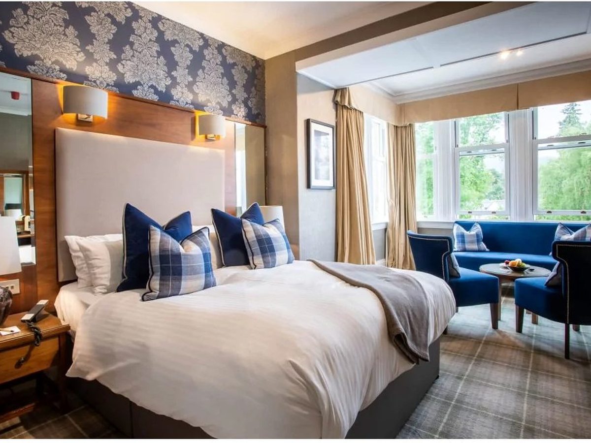 A double room with a padded headboard and blue tartan cushions, and a bay window with a blue sofa and armchairs forming a seating area to one side.