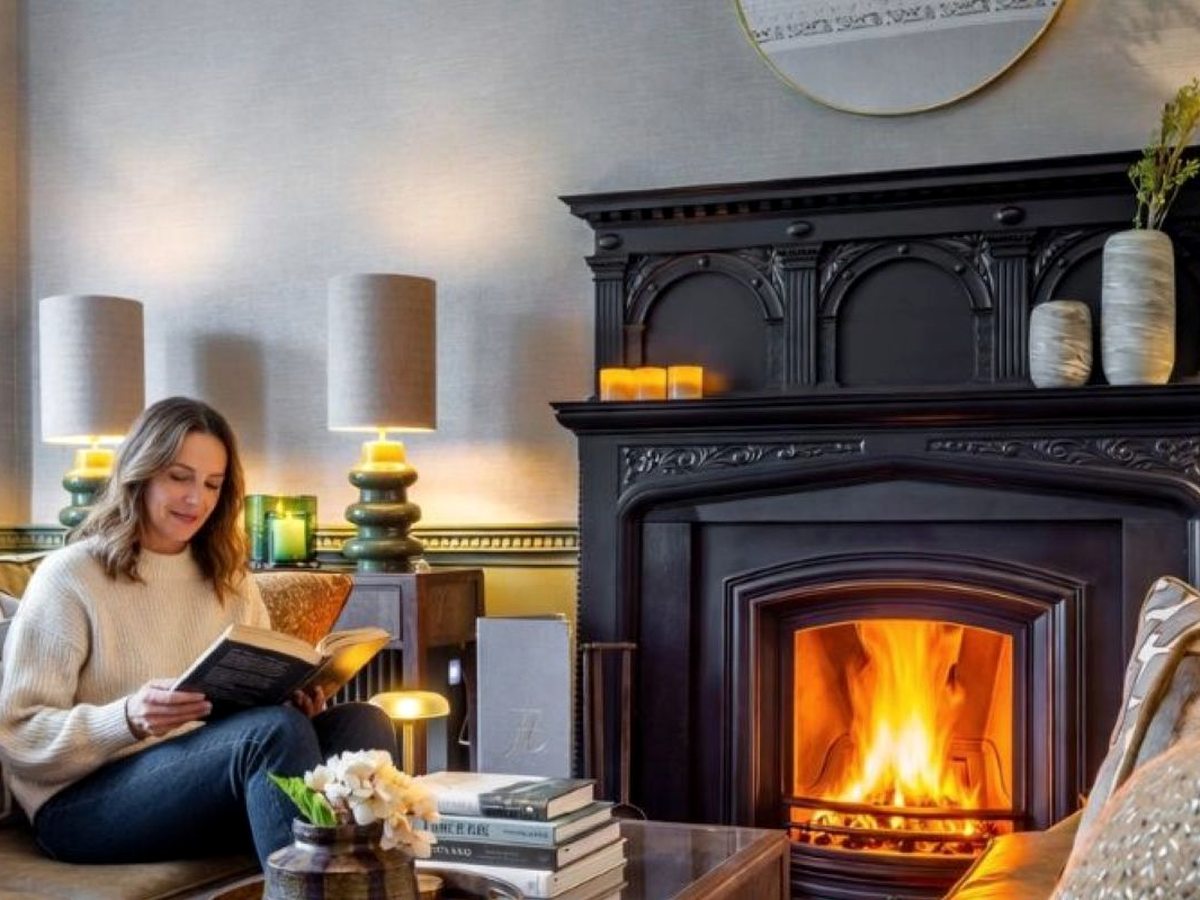 A person reading in an armchair beside a large ornate black cast-iron fireplace with a lit fire, table lamps and candles in a traditional hotel lounge.