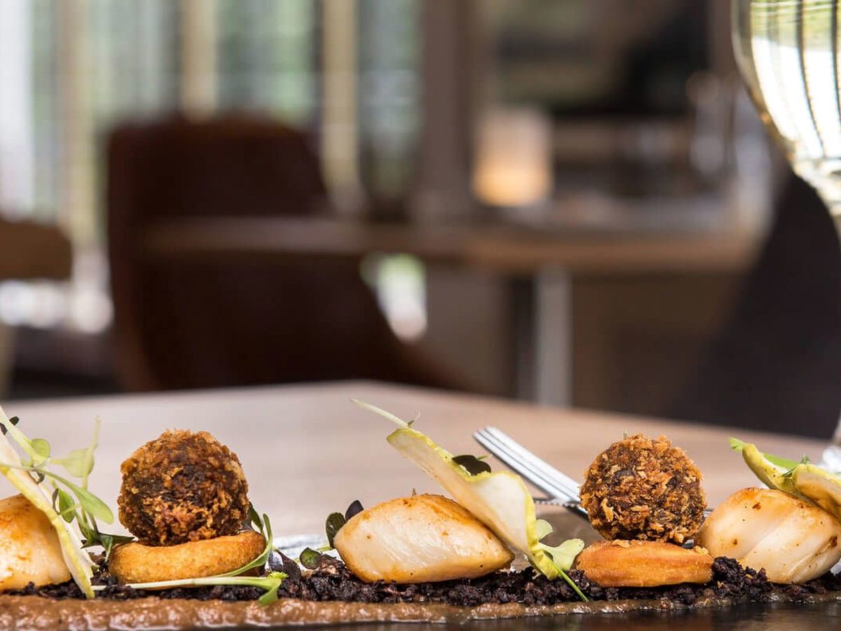 Seared scallops and breaded bonbons arranged in a line on a dark base with pea shoots, on a wooden table beside a wine glass.