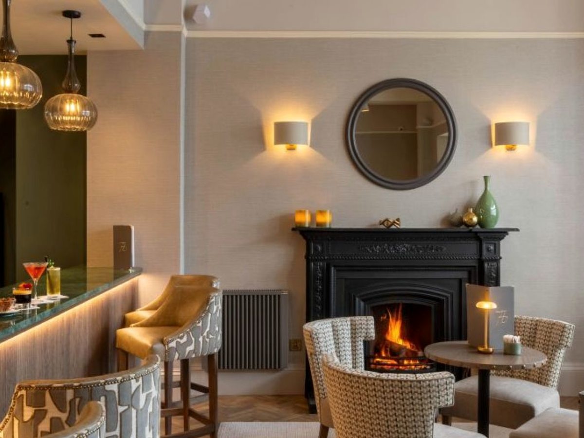 A bar counter with cocktails to one side and patterned tub chairs around a black cast-iron fireplace with a lit fire, a round mirror and wall lights above.