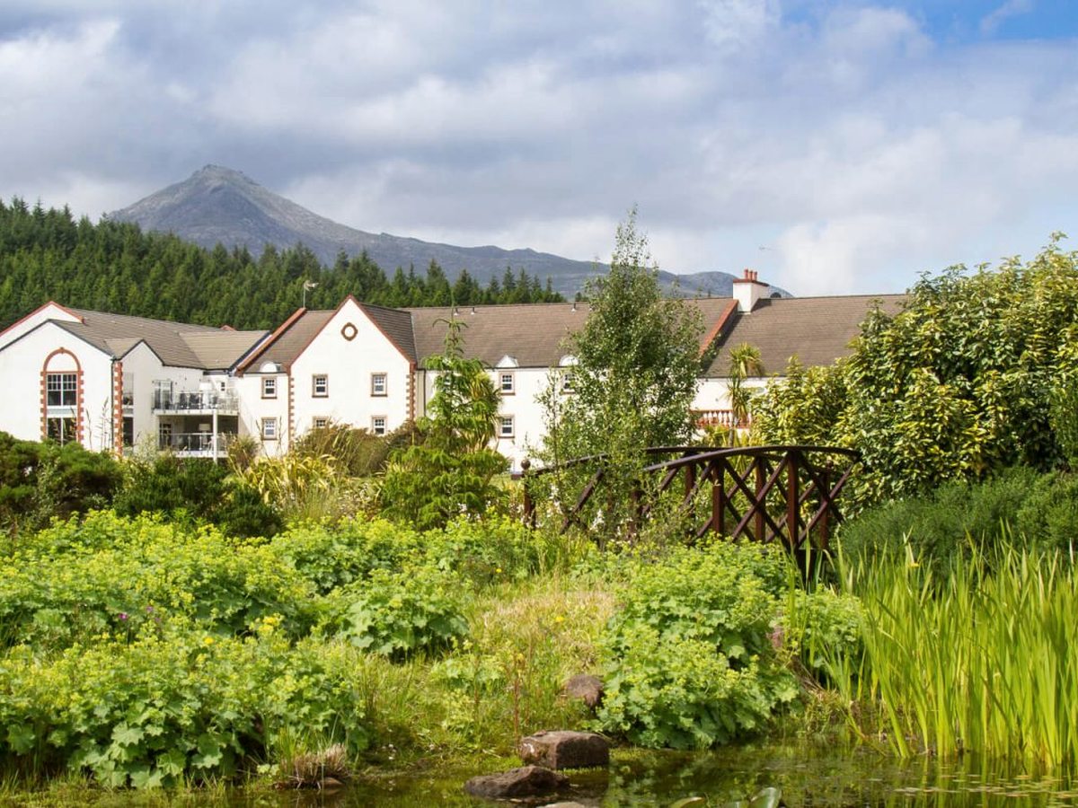 White-rendered resort buildings with balconies set below a wooded, mountain-topped hill, with a garden pond and a small wooden footbridge in front.