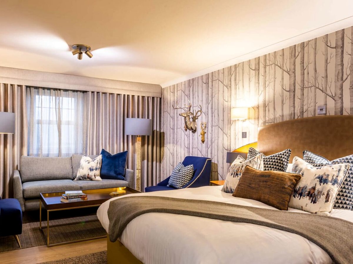 A bedroom with birch-tree wallpaper and brass stag heads, a grey sofa and blue armchairs forming a sitting area beside the bed.