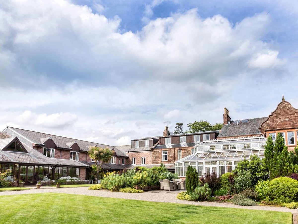 A wide manicured lawn and planted borders in front of the red-sandstone hotel and its glass conservatories under a bright cloudy sky.