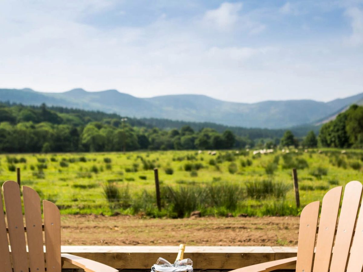 Two wooden Adirondack chairs on a decked terrace looking over a sheep pasture and trees to distant hills, with a drinks cooler between them.