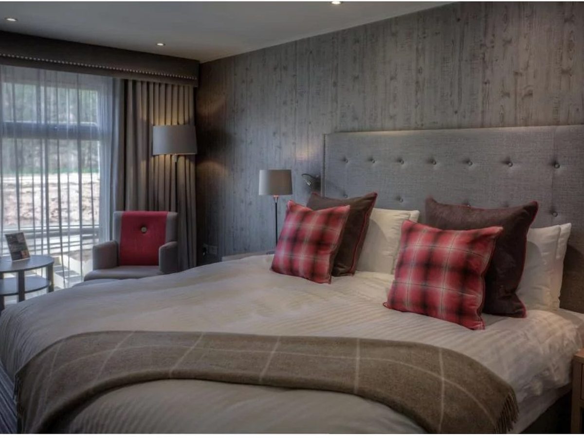 A room with a grey tufted headboard and red tartan cushions, a timber-panelled wall and glazed doors opening onto an outdoor terrace with a red armchair alongside.