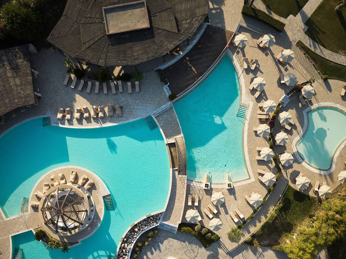 Aerial view of a large curved turquoise pool with a domed timber pavilion, loungers and umbrellas around it, and a smaller pool alongside.