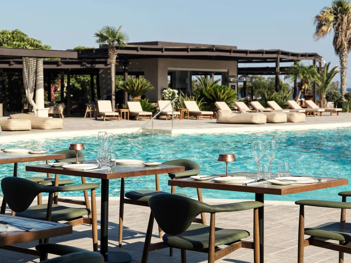 Restaurant tables set with glassware on a terrace beside a turquoise pool, with loungers and cabanas behind.