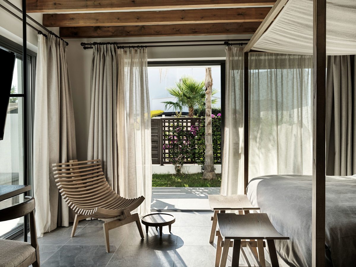 Room with a canopy bed and cane armchairs, glass doors open to a planted garden terrace.
