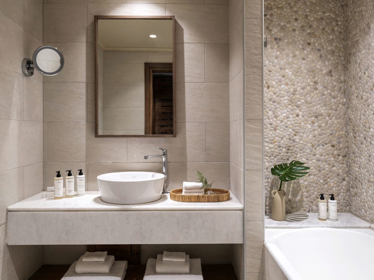 Bathroom with a round vessel basin on a stone vanity, a mirror and toiletries, and a tub beside a pebbled wall.