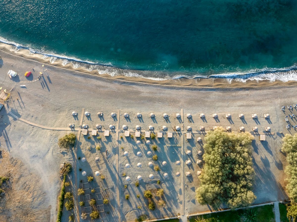 Aerial view straight down onto a beach with rows of straw parasols and the sea washing the shore.