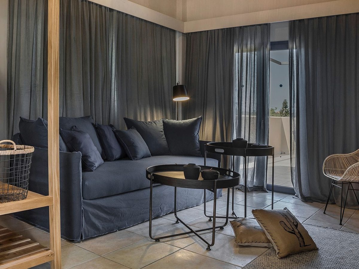 Suite lounge with a blue sofa, coffee tables, a wicker chair and open shelving beside balcony doors.