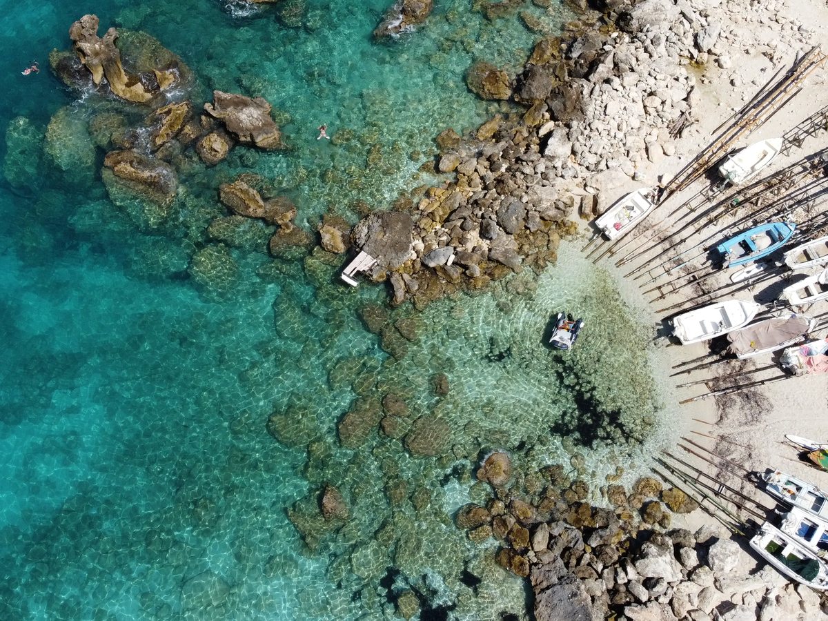 Aerial view of a rocky cove with clear turquoise water, submerged rocks, a small jetty and a few moored boats.