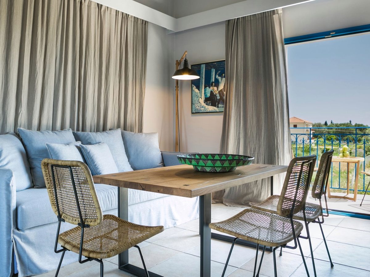 Suite with a wooden dining table and woven chairs, a blue sofa bed and a furnished balcony with a landscape view.