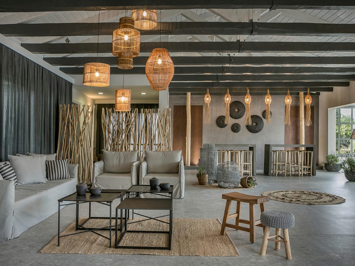Spacious indoor lounge with white sofas, armchairs and low coffee tables under rattan pendant lights and exposed beams, with a bar and high stools beyond.