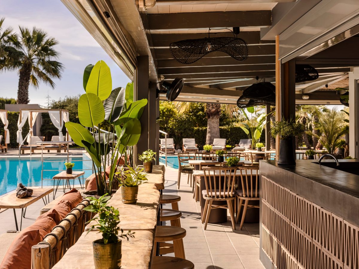 Covered poolside bar with a high wooden counter and stools, bench seating and potted tropical plants beside the pool.