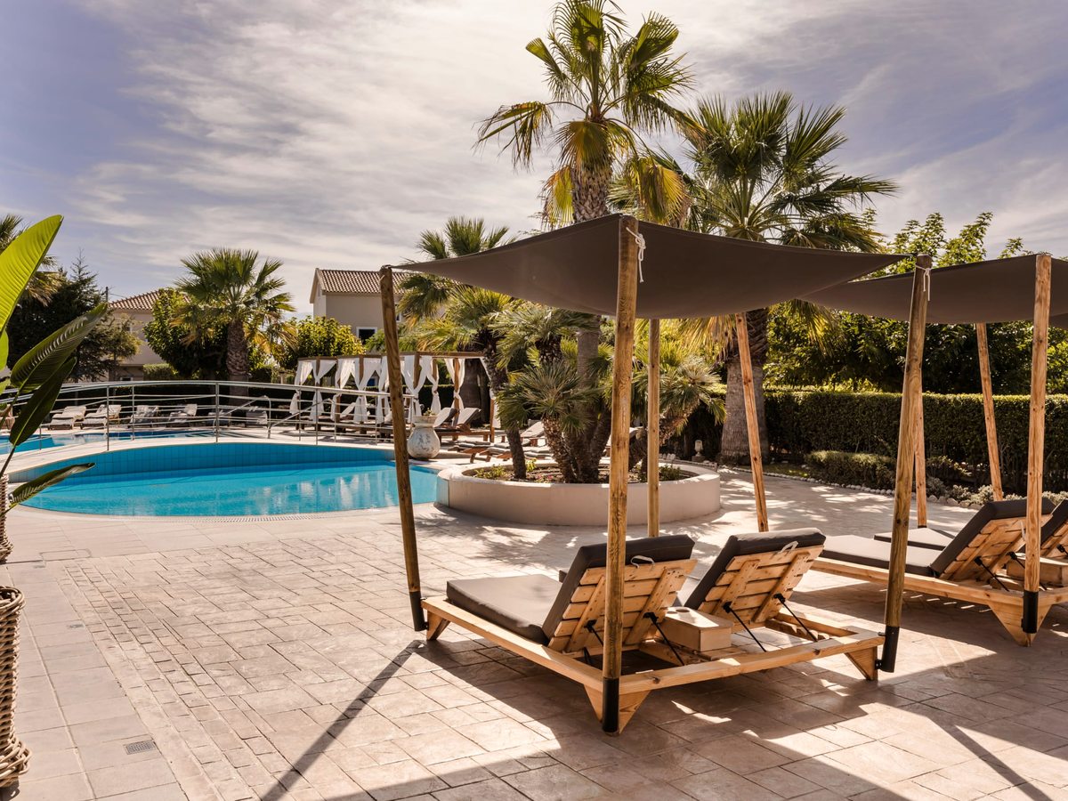 Wooden sun loungers and cabana beds under a large shade sail beside the curved pool, with palms and planted borders behind.