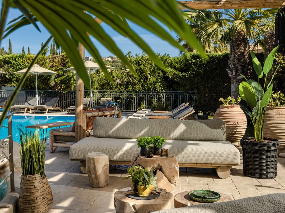 Covered garden lounge with cream cushioned sofas and low tables beside a pool railing, framed by tall palms and potted plants under a bright sky.