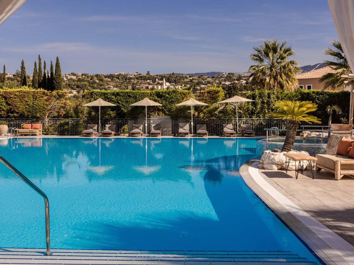 Pool steps and a handrail with sun loungers and parasols along the edge, planted borders and the village of Svoronata rising on the hillside behind.