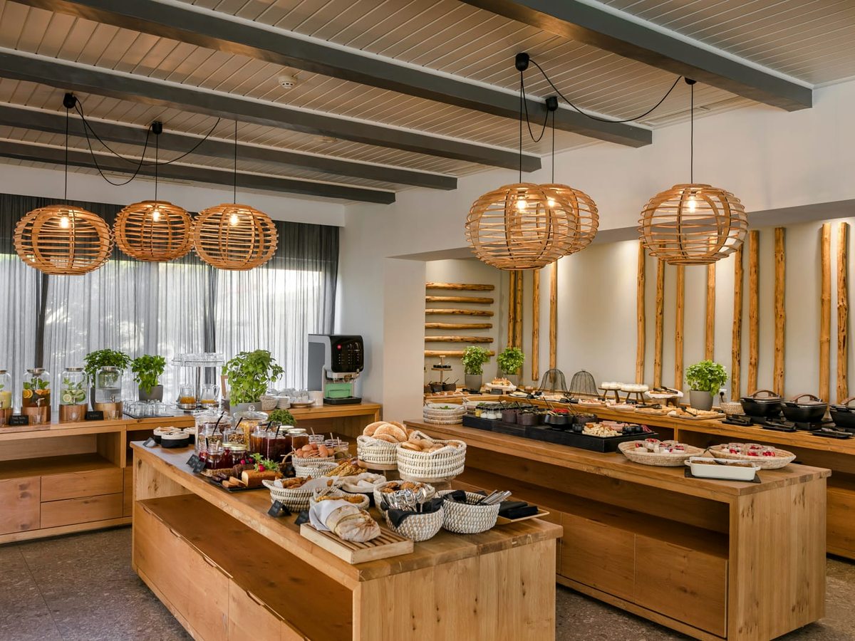 Buffet room with a central wooden serving island of hot dishes and bread baskets under rattan pendant lights and grey ceiling beams.