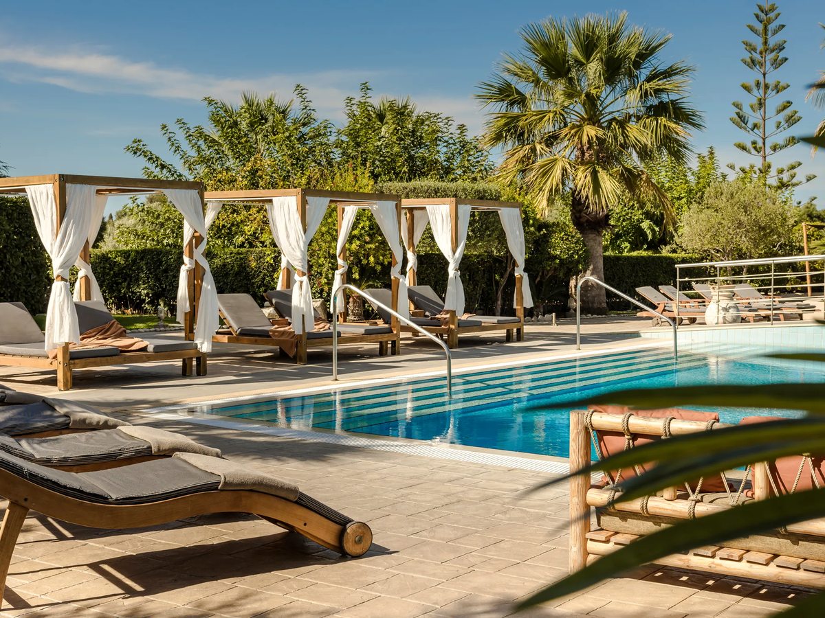 Row of white curtained cabana daybeds and sun loungers under palms beside the blue pool.