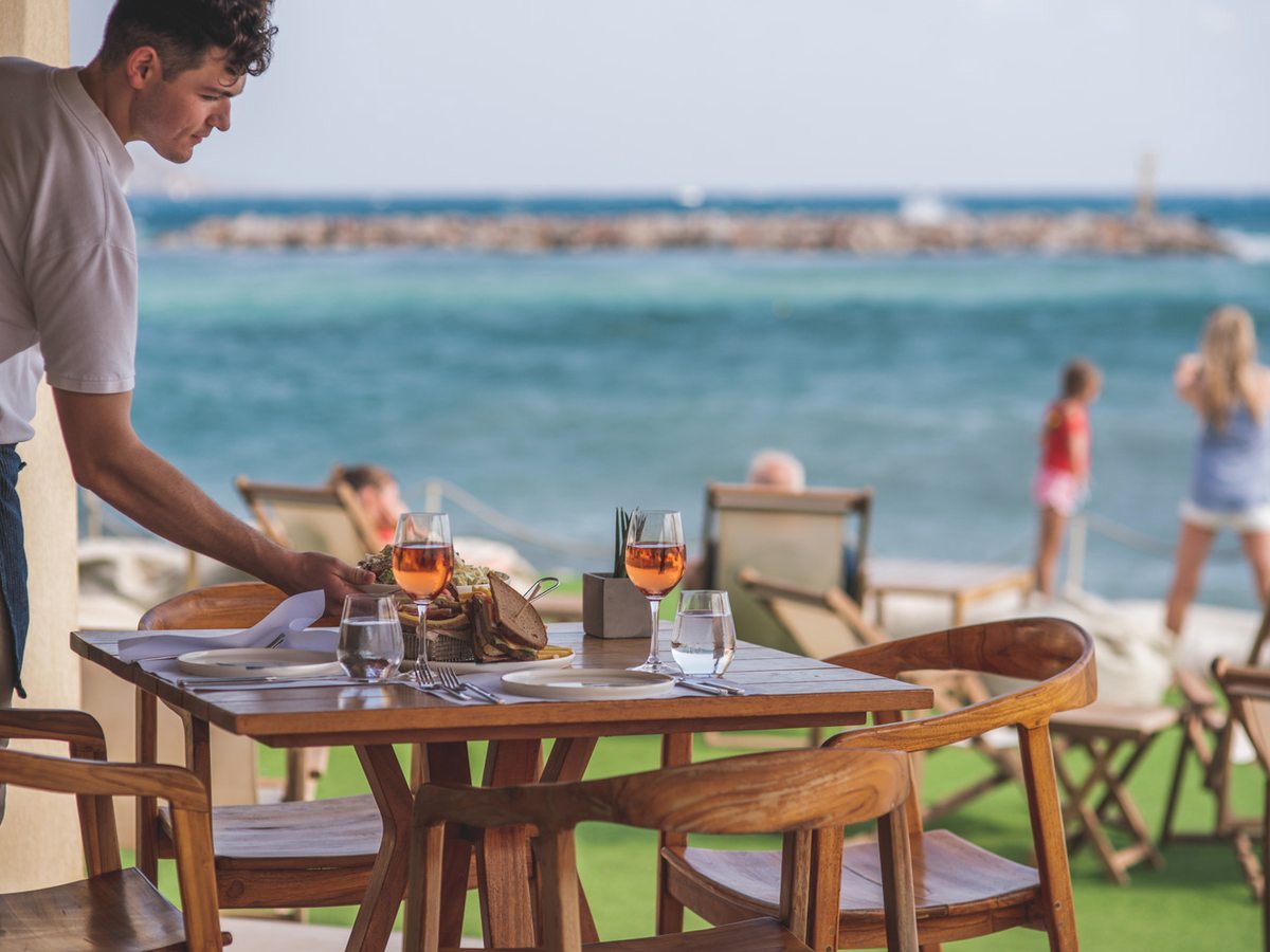 A waiter setting a wooden table on the beachfront terrace, with the sea and a breakwater behind.