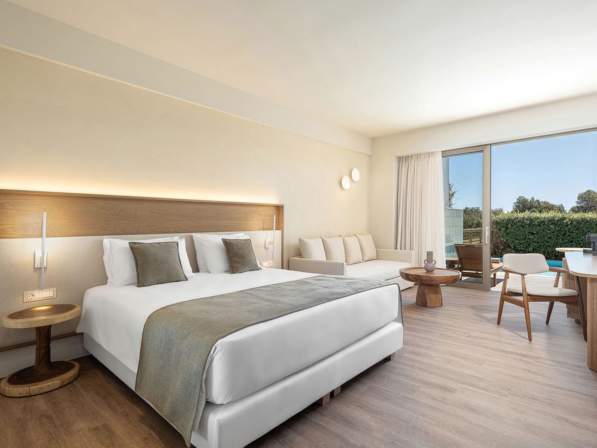 A double room with a long timber headboard, a king bed, a sofa and a desk, with a private terrace through glass doors.