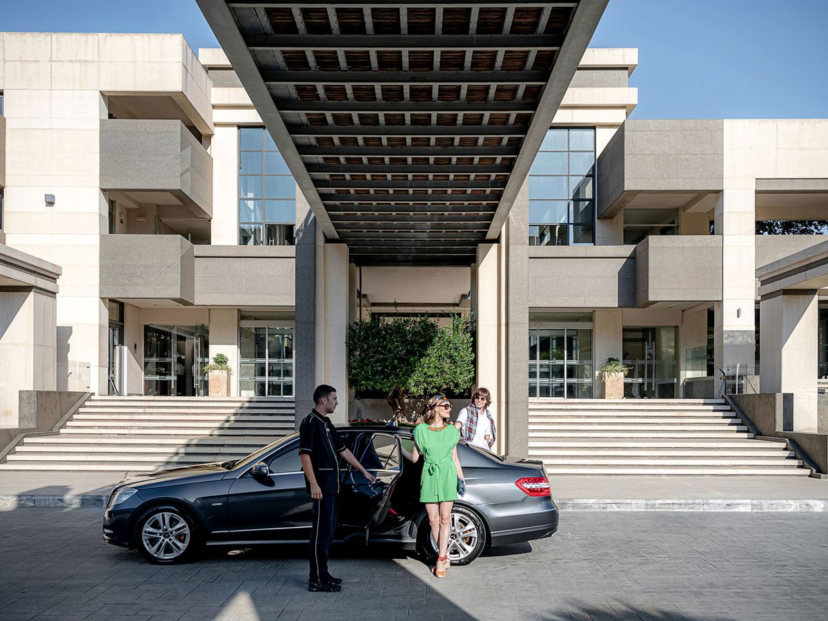 Guests arriving by car under the hotel's cantilevered stone entrance canopy, with wide steps up to the glass doors.