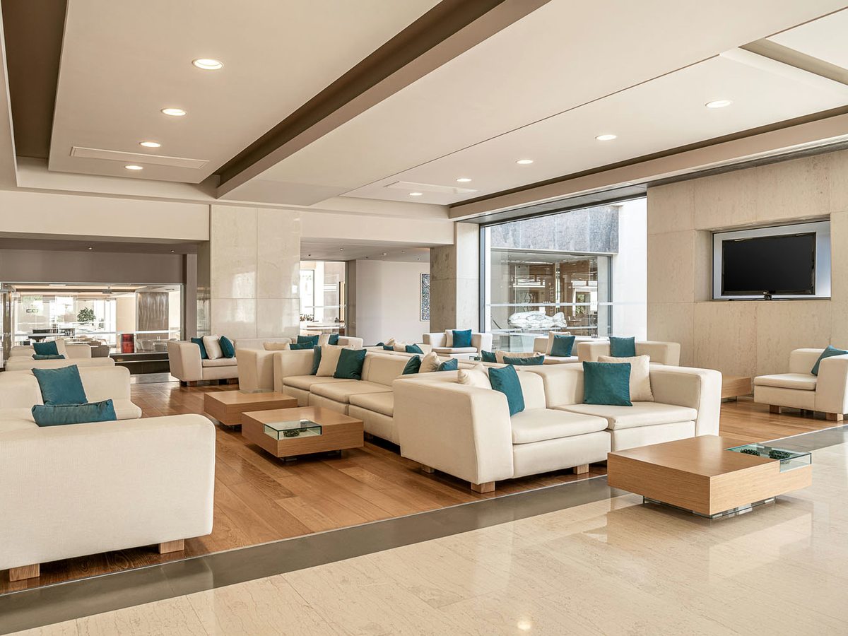 A lounge of white sofas with turquoise cushions and glass coffee tables on a wood-effect floor beneath a wall-mounted screen.