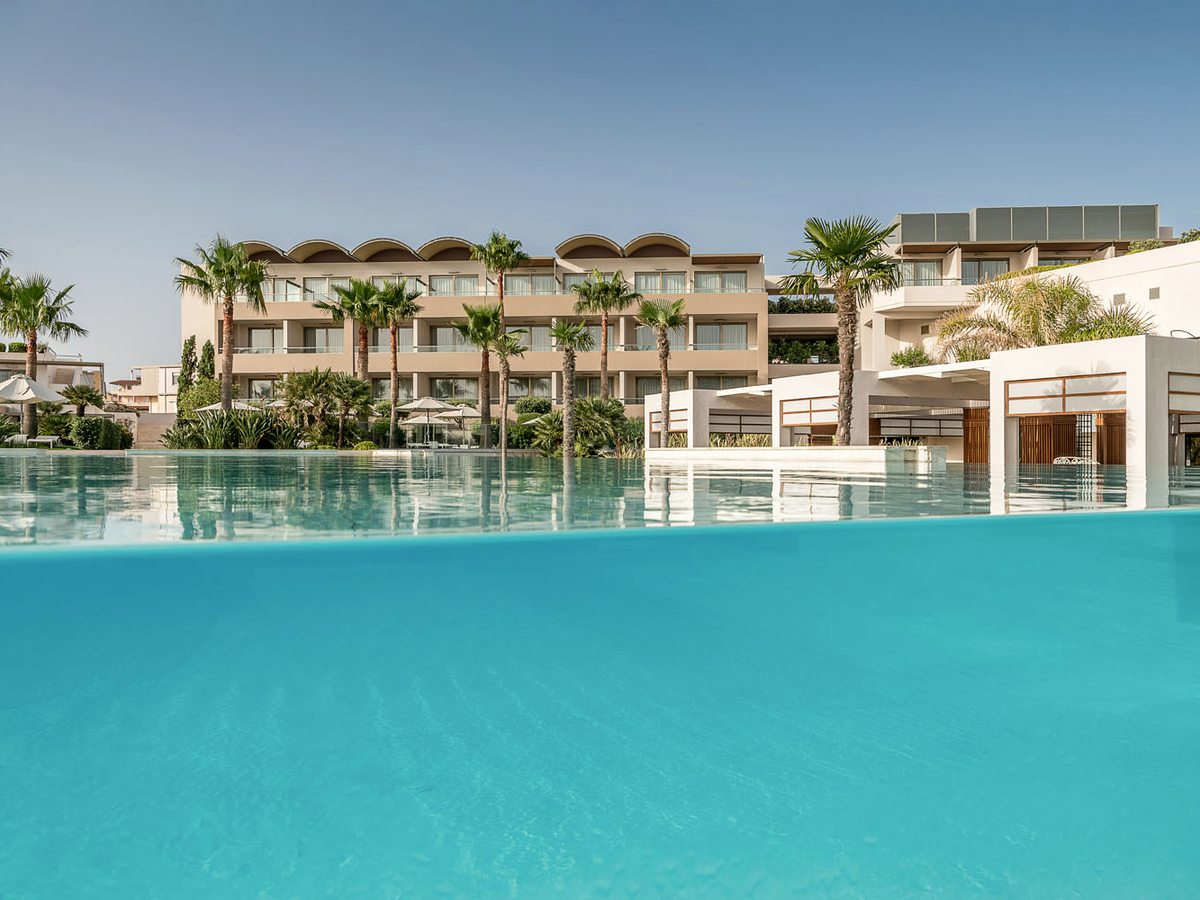 The large central pool seen from water level, its clear glass side revealing the resort buildings and palms behind.