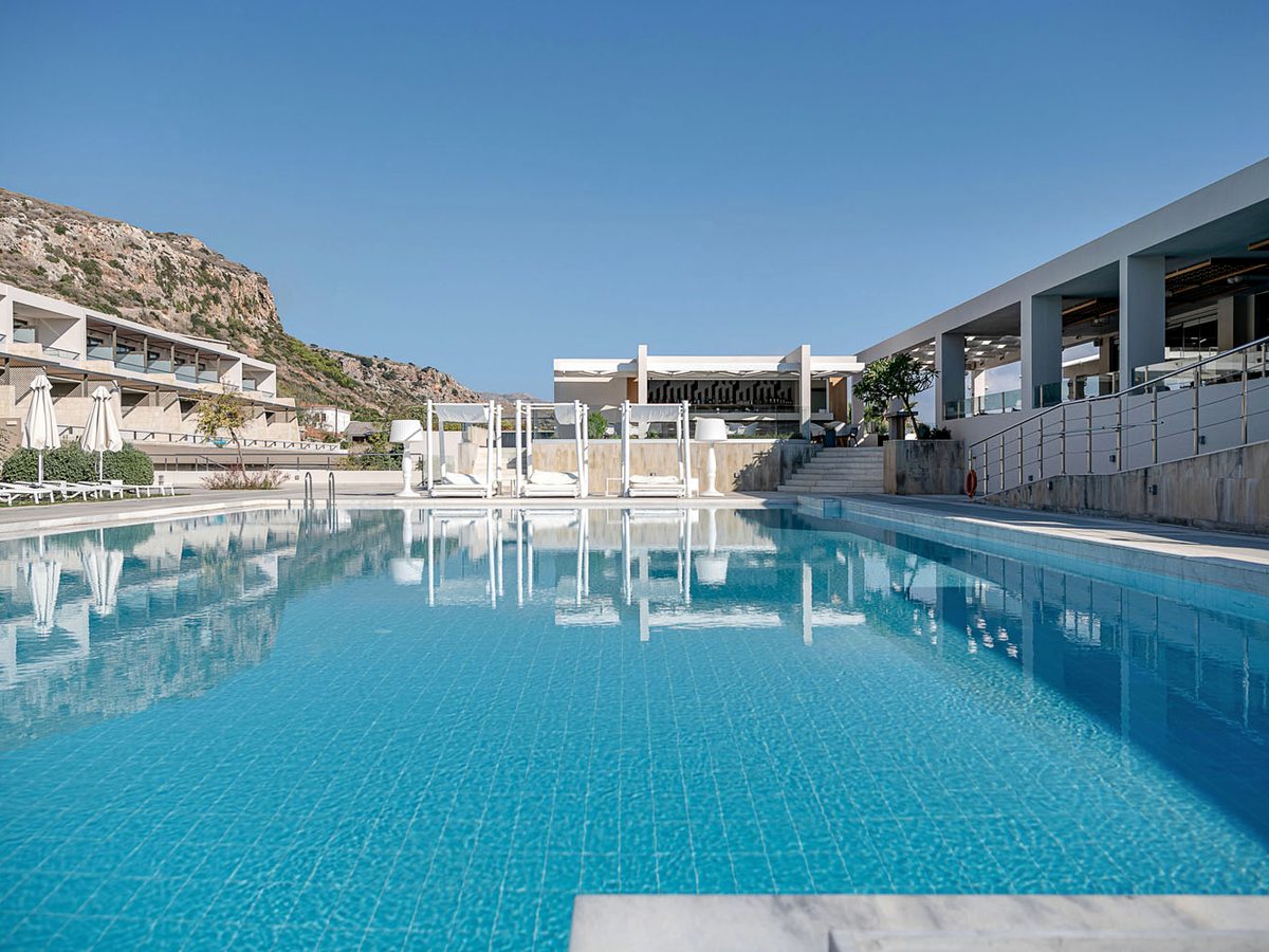 A long rectangular pool with sun loungers and parasols along the deck and a covered terrace beside the rooms.