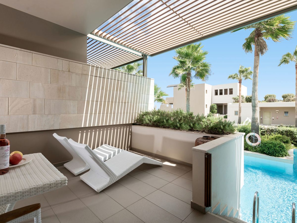 A room terrace with loungers under a slatted pergola and steps down into the shared pool beyond.