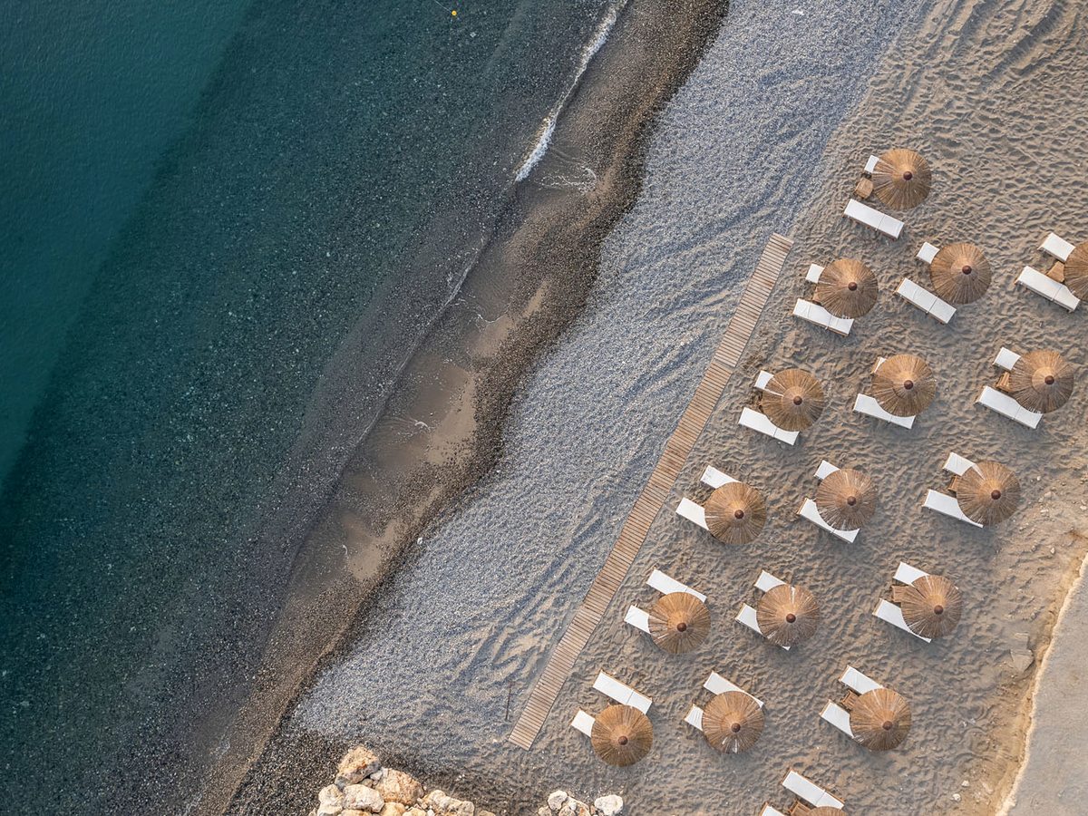 An aerial view of straw parasols and white sunbeds in rows along the pebbly shore and clear shallows.