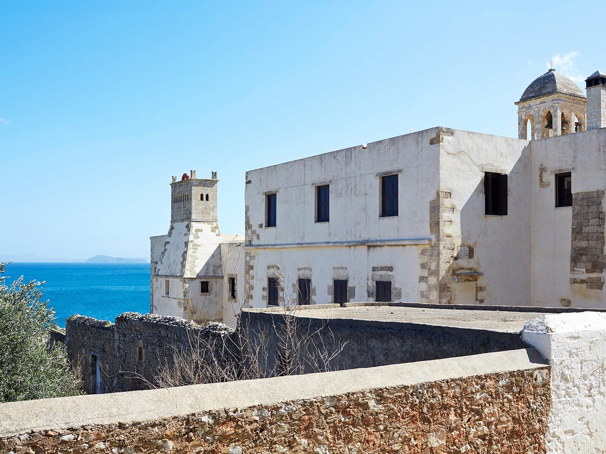A whitewashed monastery complex behind a stone wall on the coast, under a clear blue sky.
