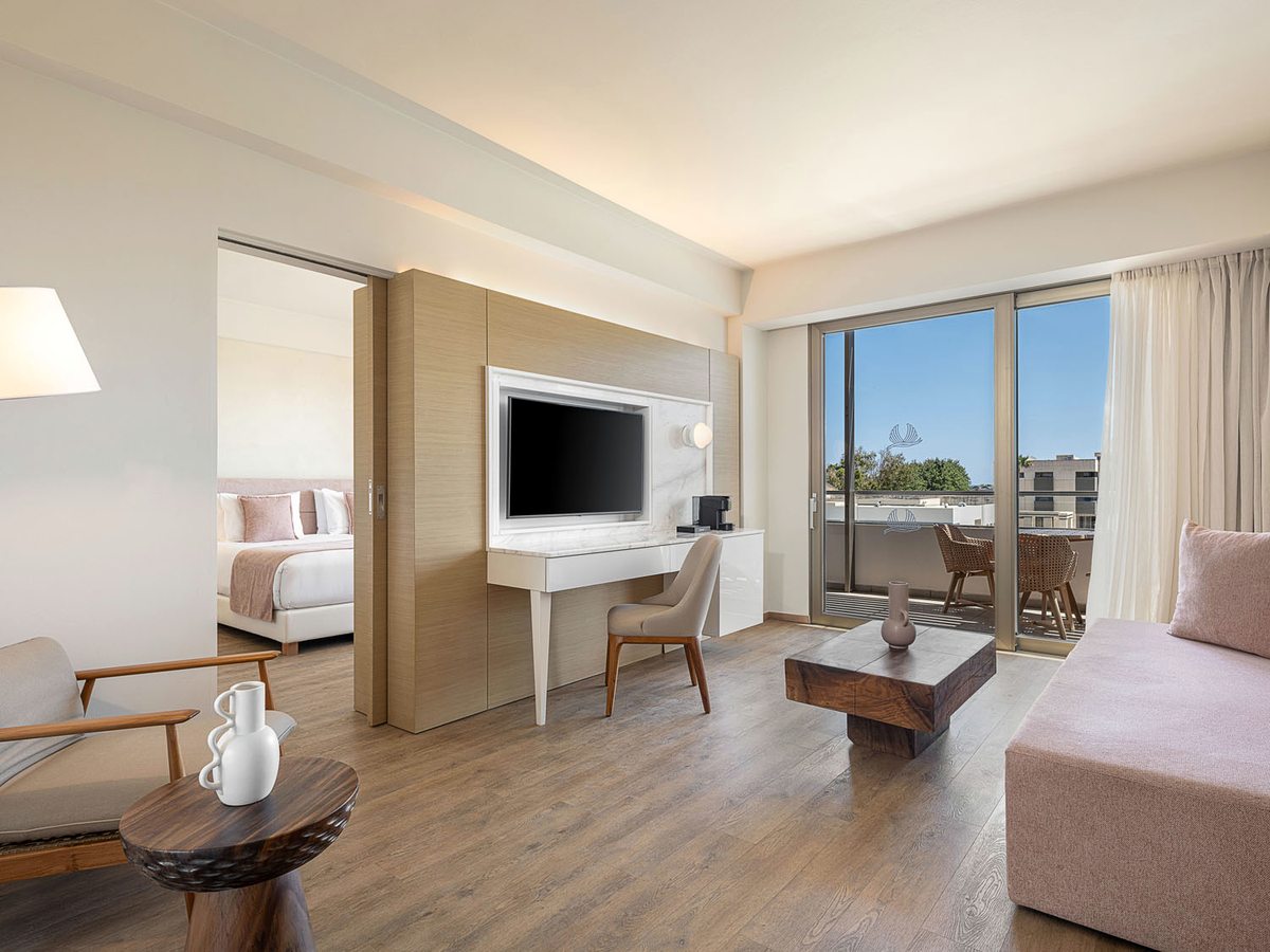 An open-plan suite with a lounge, coffee table, desk and wall-mounted screen, and a sleeping area beyond a sliding partition.