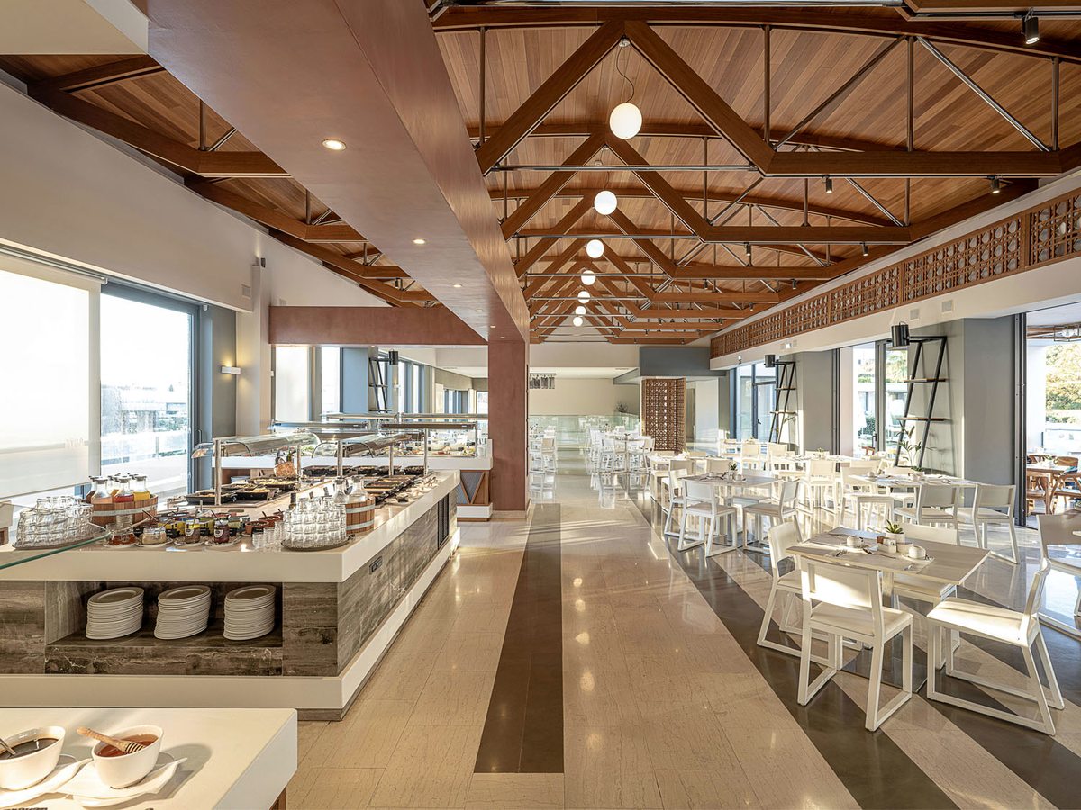 A buffet restaurant with a long serving counter of hot dishes under a timber-truss roof, and white tables and chairs down one side.
