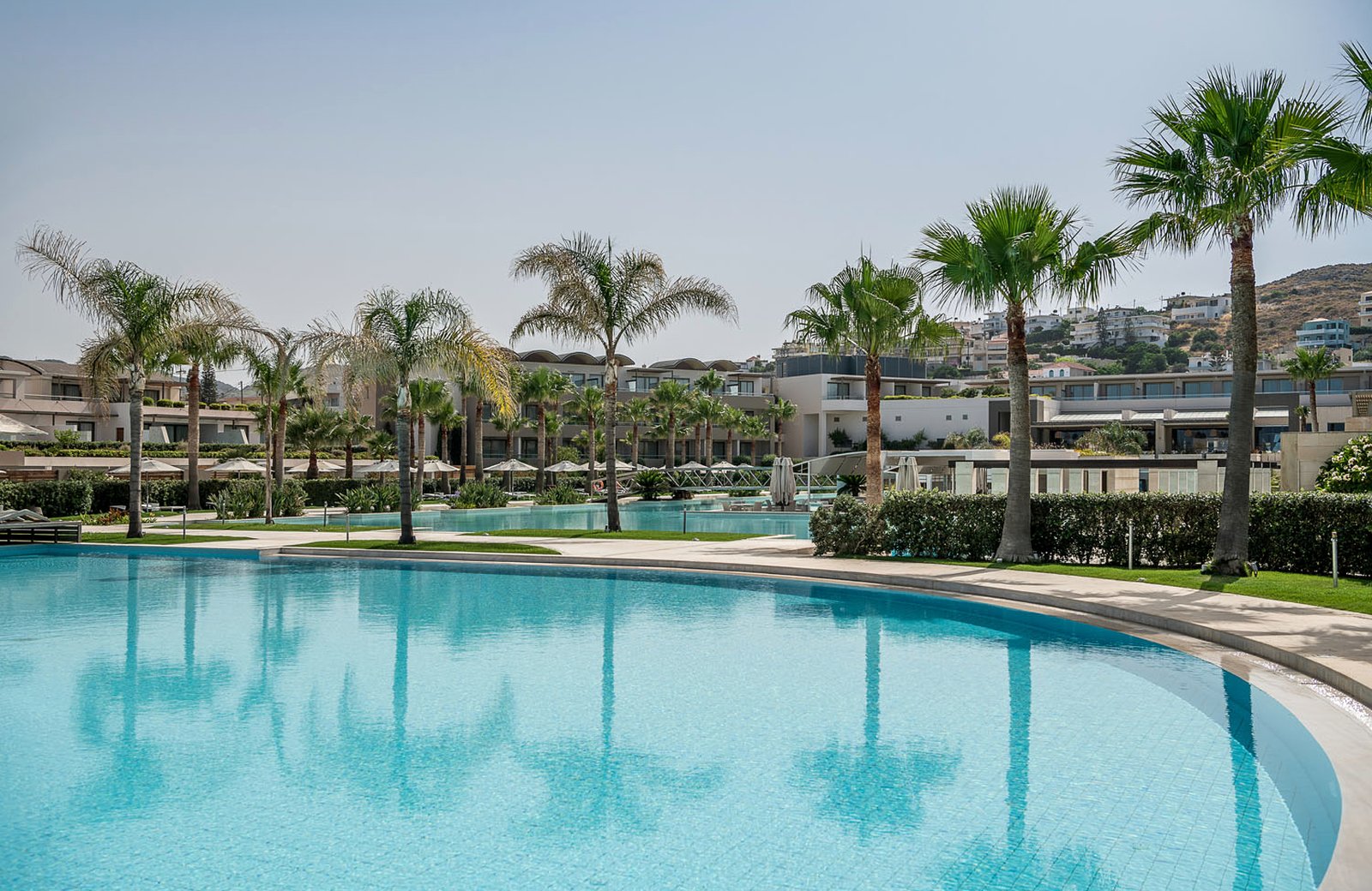 The large main pool at Avra Imperial, ringed by palms with the resort's low-rise buildings and the hillside behind Kolymvari beyond.