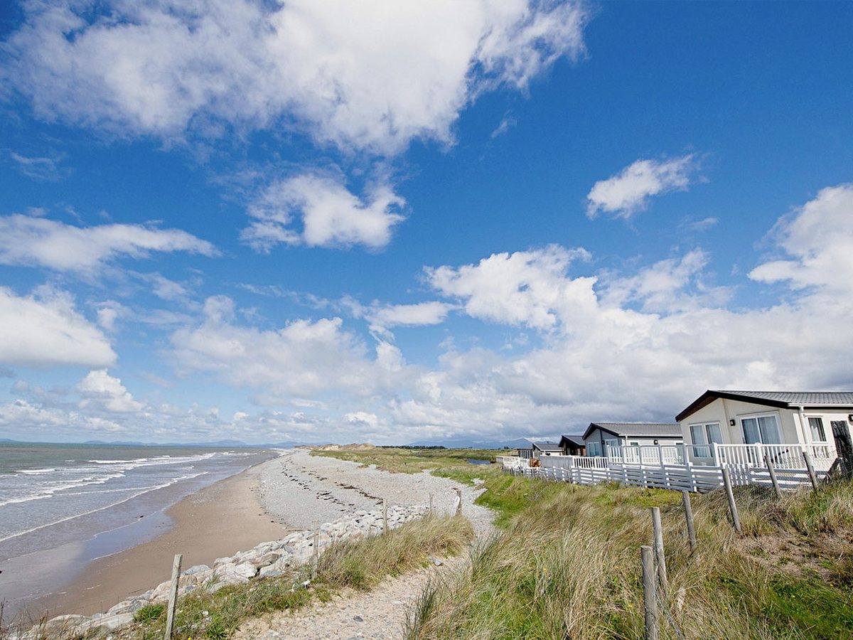 A wide sandy beach curving along the coast, with grassy dunes and white caravans to one side under a blue sky.