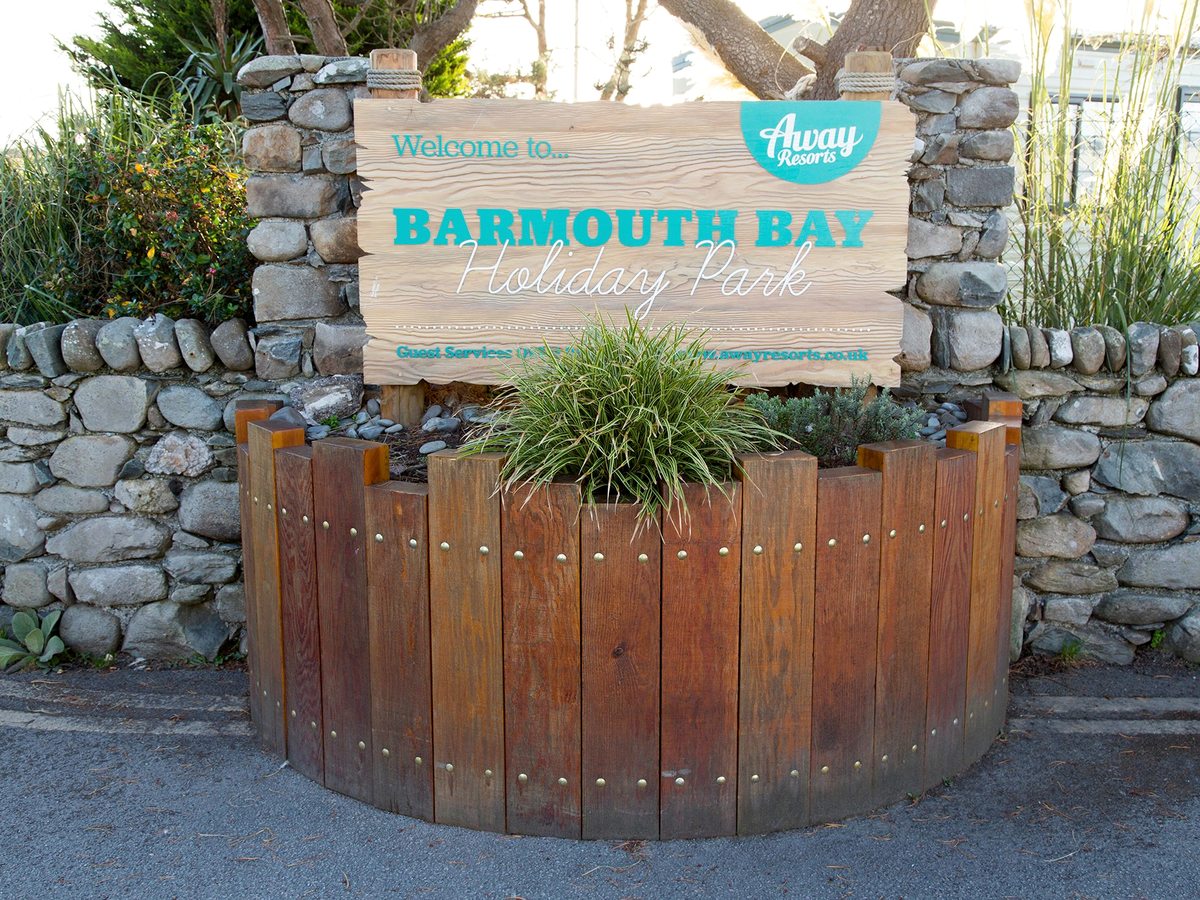 A low stone-walled entrance with a wooden planter and a painted Welcome to Barmouth Bay Holiday Park sign.