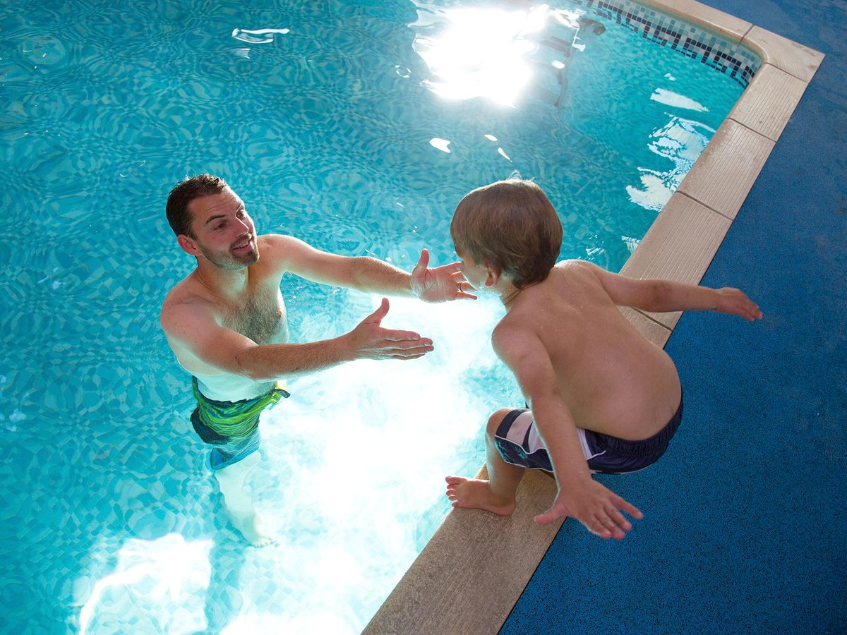 A man and a young boy at the tiled edge of a sunlit indoor pool.