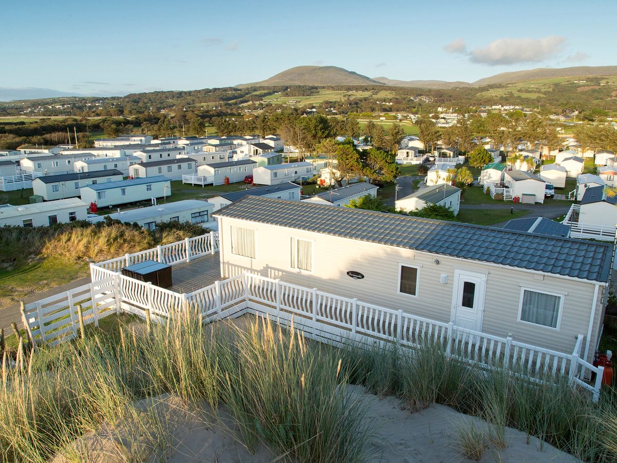 An aerial view of white caravans among grassy dunes, with green hills and mountains along the horizon.