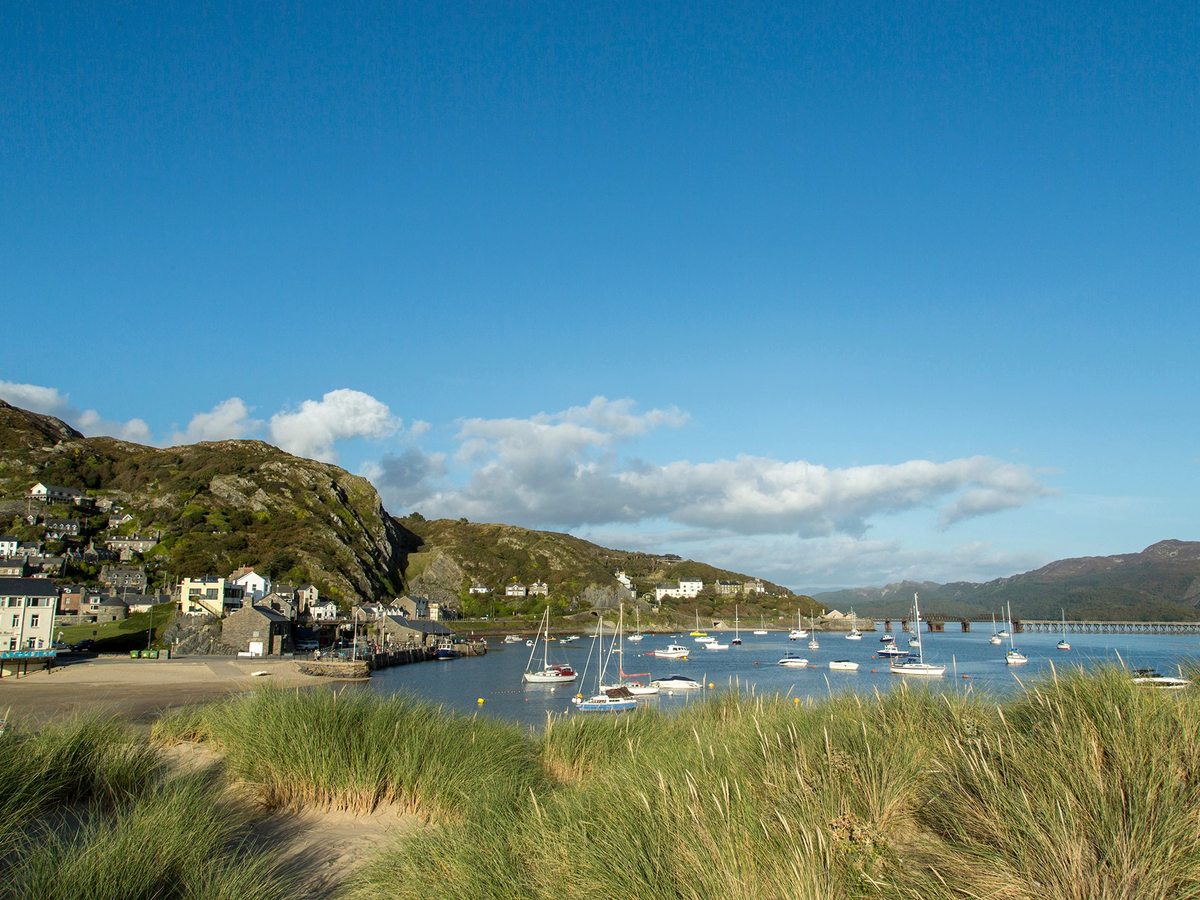 A small harbour with moored sailing boats, waterfront houses and a rocky hillside behind, seen across dune grass.