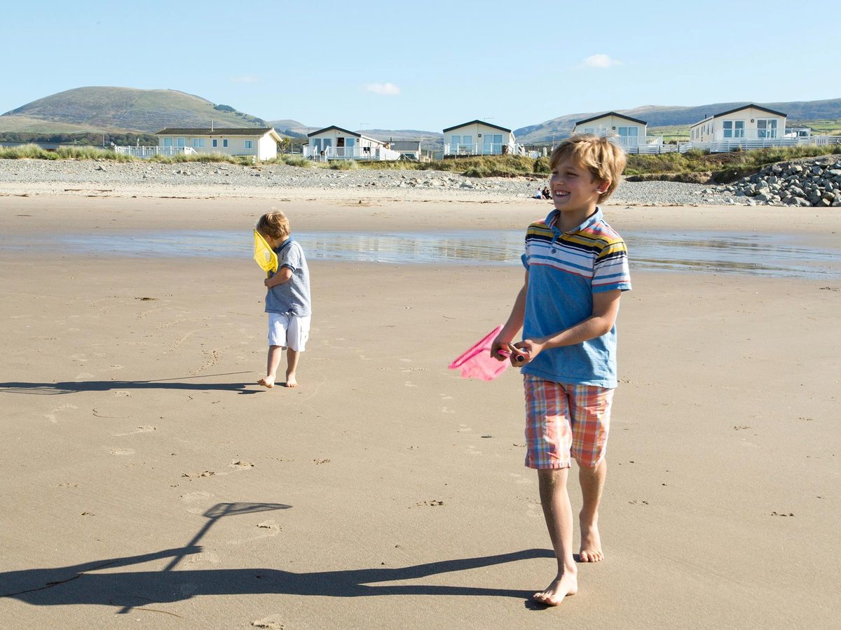 Two children walking across wide wet sand at low tide, with caravans and green mountains in the distance.