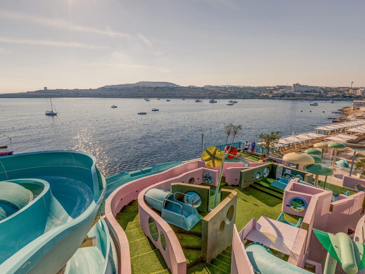 An outdoor children's water-play area with pastel play structures and twisting blue water slides on artificial grass, set above a bay dotted with small boats.