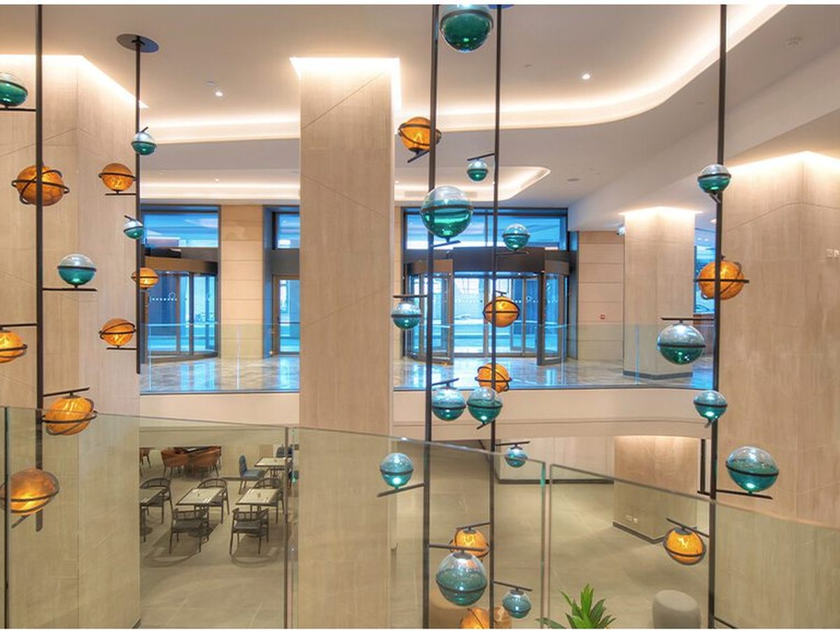 A double-height lobby atrium with stone-clad columns, a glass balustrade around the upper level and a cascade of blue and amber glass pendant lights hanging through the void over a polished floor.