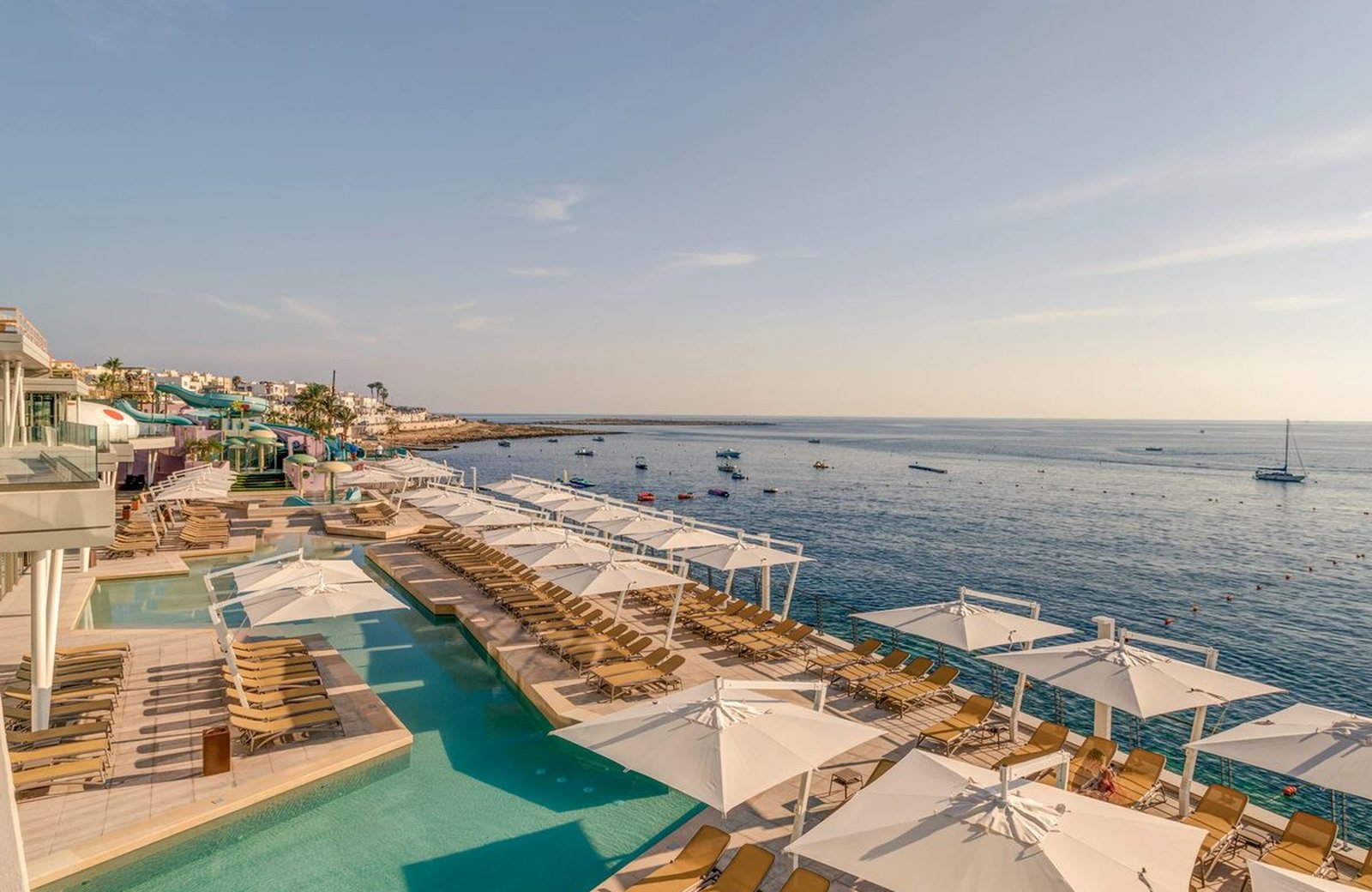 The panoramic lido at AX ODYCY, a long outdoor pool edged with sun loungers and white parasols on a terrace at the water's edge, looking out over Qawra Bay.