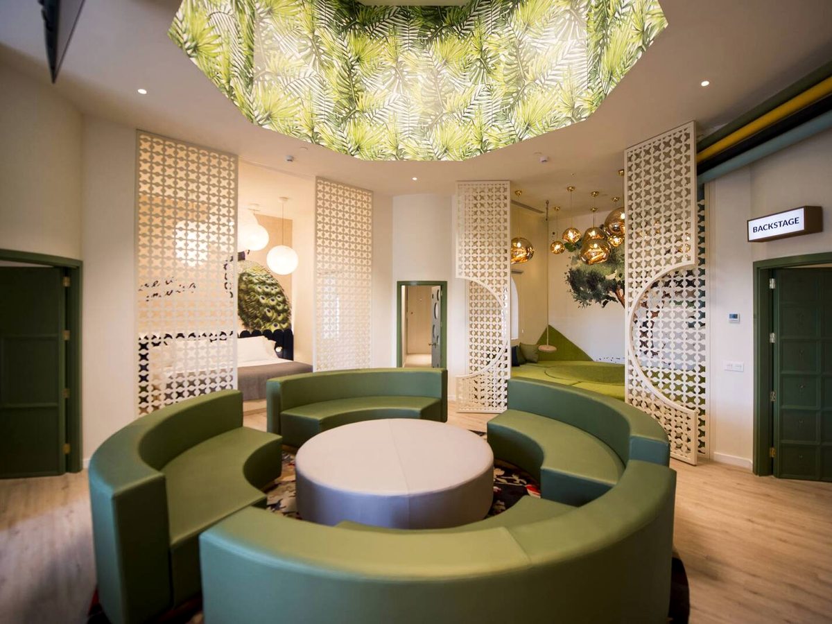 A suite's central lounge with a green circular sofa, a leaf-print illuminated ceiling and white lattice screens, a themed bedroom visible beyond.